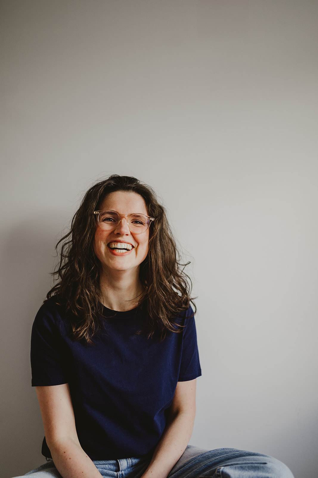 a woman in a dark blue T-shirt and jeans sitting cross-legged in front of a white wall and smiling broadly